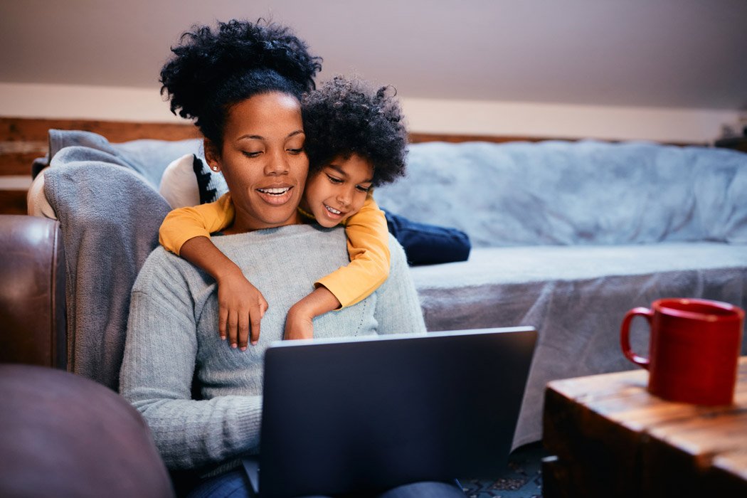 Young boy smiling and holding his mom while she looks at her laptop.
